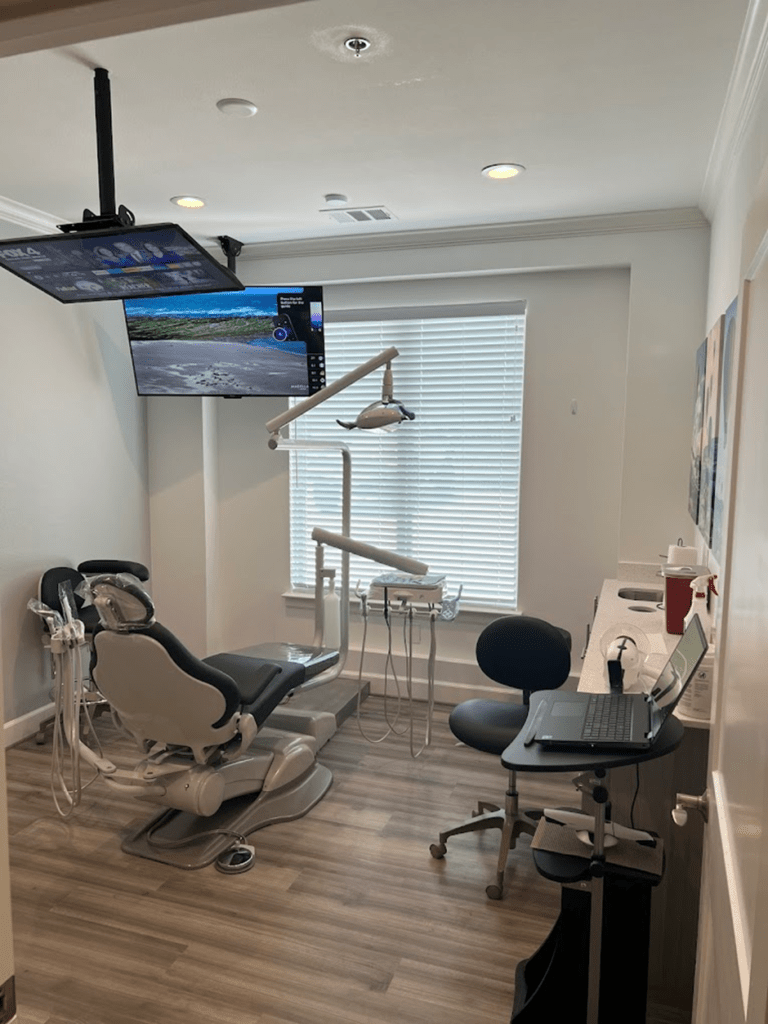 A modern dental office in Frisco with an adjustable chair, overhead light, countertop with dental instruments, and two stools. Two screens are mounted on the ceiling, one displaying a nature scene with sea creatures, and a large window with blinds provides natural light. It's truly the best dental clinic in Frisco.