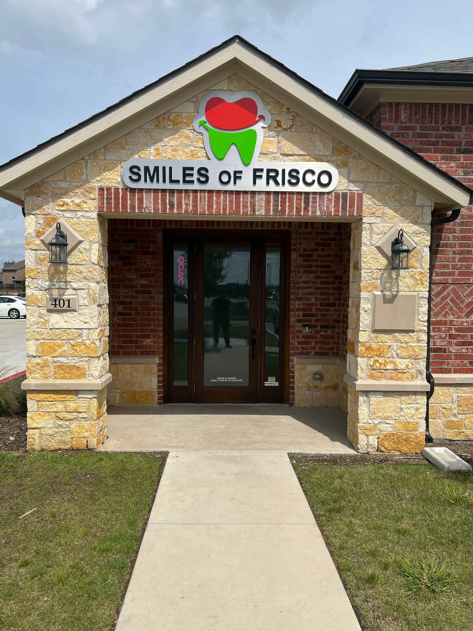 An entrance to the best dental clinic in Frisco, "Smiles of Frisco," boasts a stone exterior with a brick accent. The dark wooden door is flanked by two wall-mounted lanterns, while the sign above features a green tooth and red smiling lips logo.